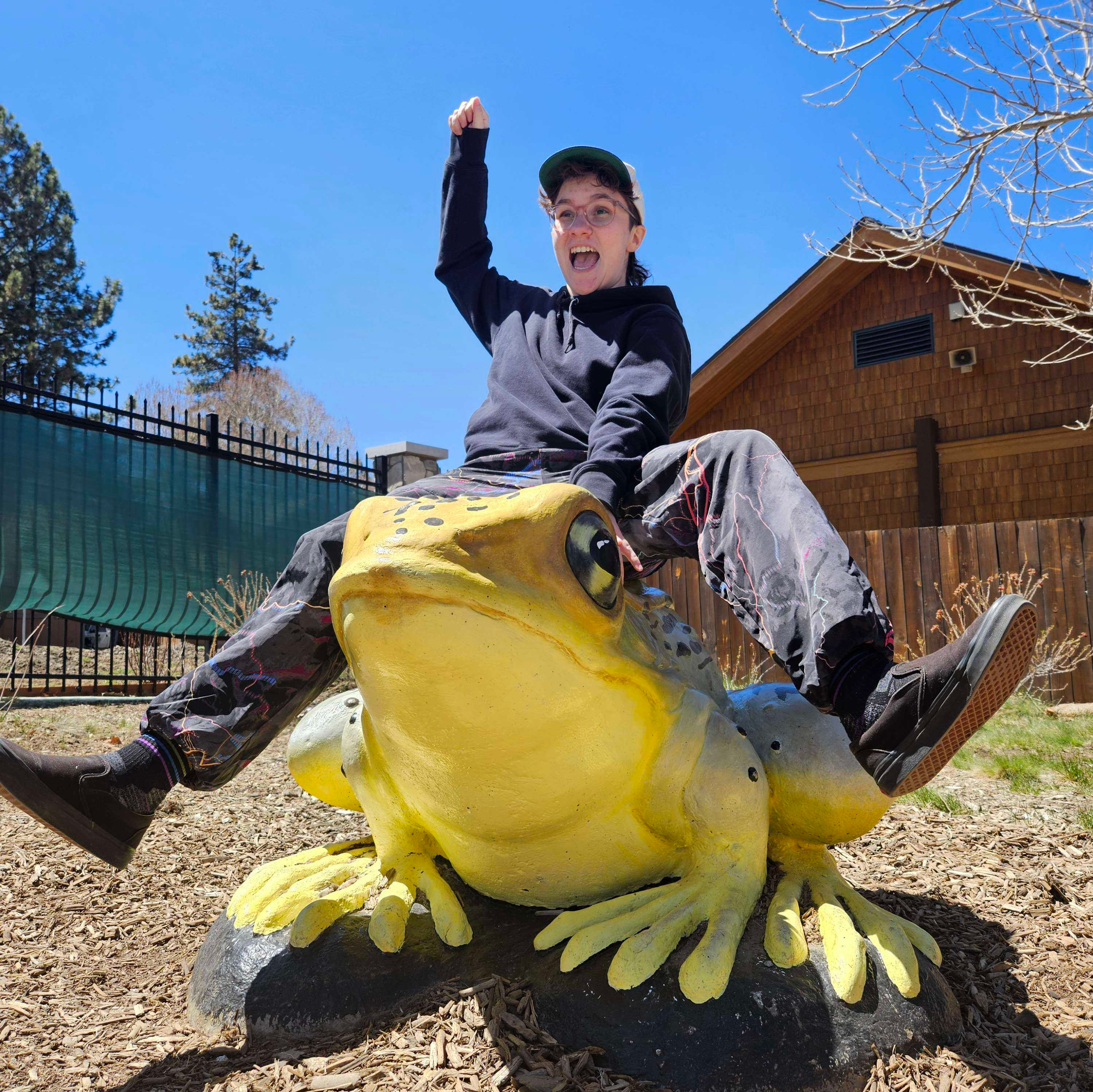 a cropped photo of alex pretending to ride a frog statue at a zoo. they have on a black hoodie, a hat, and patterened colorful pants. they have their hand up as if they are a rodeo cowboy.